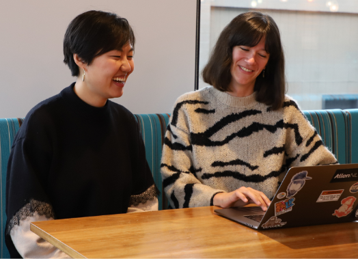 A person with dark short hair and a dark sweater is ecstatically smiling at another person with shoulder-length dark hair and a zebra sweater gesturing towards a laptop.
