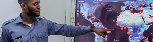 A black person with a concentrated face wearing a blue button up shirt points at a large monitor displaying a map of the world that has red dots on it.