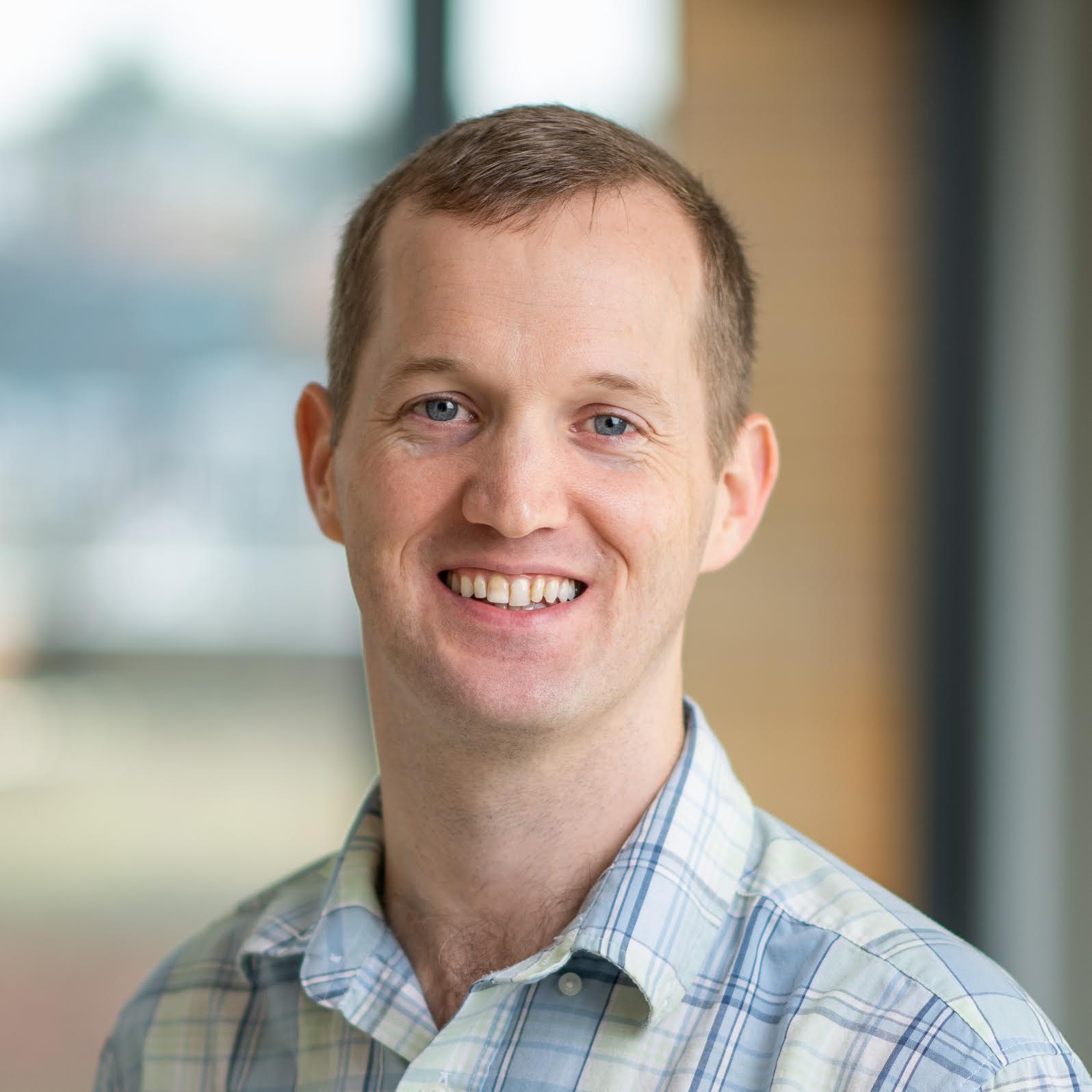 Smiling person with short hear and a blue plaid shirt