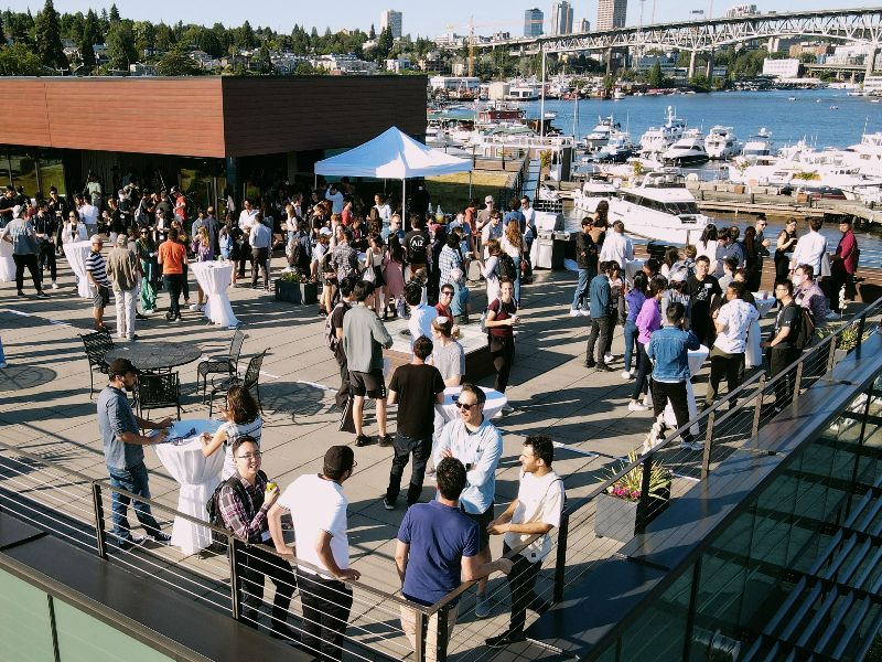 A large group of people socialize together on a rooftop deck in the sun with Lake Union and some of the North Seattle skyline in the background.