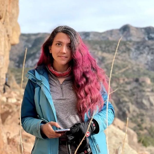A photo of Kiana Ehsani, a woman with long wavy pink hair wearing a blue jacket standing in a mountainous landscape.