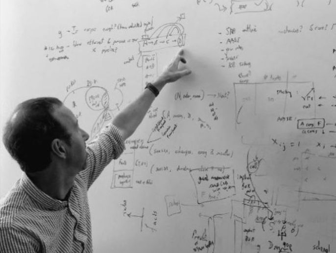 A photograph of AI2 interim CEO Peter Clark standing at a white board with lots of equations written on it, pointing to one.